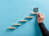 Fototapeta  - Male hand arranges a wooden block staircase with target icon. Achieving goals and objectives or goal setting