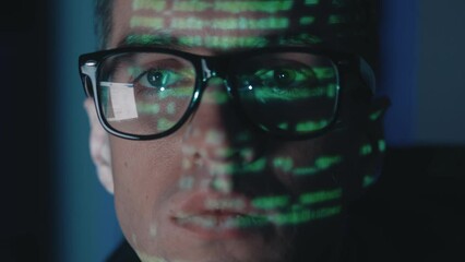 Close-up face portrait IT hacker programmer developer in glasses working computer, cyber attack on server at night. Green program code reflected on face. Male virtual spy hacking software in dark room