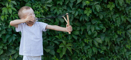 a boy in a T-shirt on the background of a hedge aims a slingshot. banner with space for text