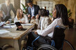 © Lomb - Office coworkers in the meeting room - main focus on African American woman with disabilities sitting on the wheelchair - business and disability lifestyle concept
