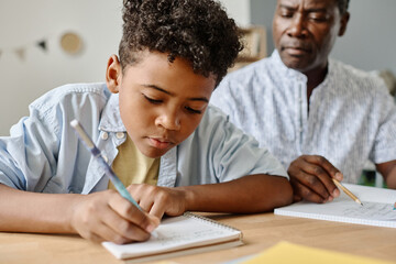 Wall Mural - African little boy making notes in his notebook at table and doing homework with teacher