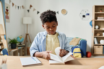 Wall Mural - African little boy turning the pages of his book at the table while doing his homework at home