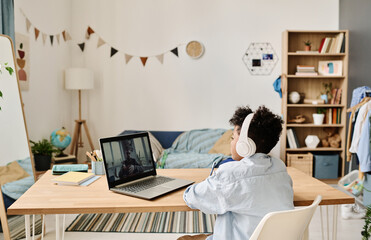 Wall Mural - Rear view of African schoolboy in headphones having online video call on laptop with his teacher at table in the room