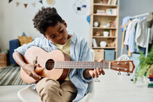 African Boy Sitting On Chair And Concentrating On His Play In Guitar, He Remembering The Notes During Home Lesson