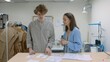 © stanis88 - 4K. A young female seamstress works with a client in a sewing workshop. They are looking at sketches of dresses