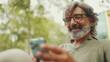 © Andrii Nekrasov - Positive middle-aged man with gray hair and beard wearing casual clothes sits on bench and uses mobile phone. Mature gentleman in eyeglasses writes message on smartphone