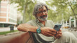 © Andrii Nekrasov - Positive middle-aged man with gray hair and beard wearing casual clothes sits on bench and uses mobile phone. Mature gentleman in eyeglasses writes message on smartphone