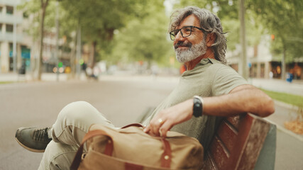 Positive middle-aged man with gray hair and beard wearing casual clothes sits on bench. Mature gentleman in eyeglasses is resting on bench
