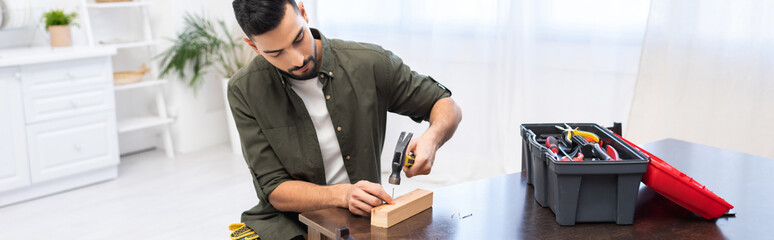 Arabian man holding hammer near nail on board near tools on table at home, banner