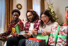 African-looking Guy Holds Book In Hands And Discusses What He Read With Friend. Friends Are Sitting On Couch And Next To Them Is Smiling Girl Of Asian Appearance, Who Is Unpacking Christmas Present.