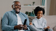 © Yuliia - Close-up happy african american father and daughter laughing watching funny tv show movie relaxing on couch man holding remote control showing thumb up gesture of approval dad and kid girl having fun