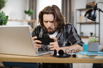 Wall Mural - Attractive bearded man playing video games on his smart phone during break in bright modern office or home. Concept of rest after hard work.