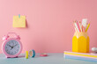 Back to school concept. Photo of school supplies on blue desk pen holder alarm clock stack of copybooks adhesive tape stapler and sticky note paper attached to pastel pink wall