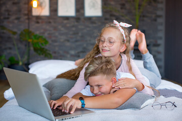 Fair-haired children - a boy and a girl study and play at a laptop, lying on the bed