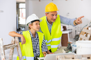 Wall Mural - Young boy and his father wearing protective helmets and yellow vests, discussing work plan in new apartment, using laptop. Man making pointing finger gesture.