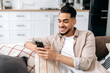 © Kateryna - The concept of using gadgets. Positive handsome indian or arabian guy, sit in a modern living room on comfortable sofa, uses his smartphone, texting message, browsing social media, surfing web, smiles