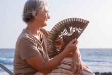 Portrait Of Beautiful Older Woman Sitting On The Beach At Sunset While Blowing Air With A Fan - Relaxed Elderly Lady Enjoys Vacation And Freedom