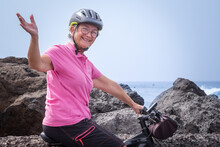 Active Happy Senior Woman With Helmet Running On Her Electric Bicycle Along The Sea Looking At Camera. Freedom And Vacations Concept