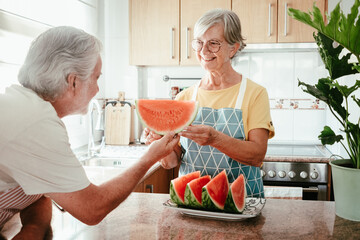 Wall Mural - Smiling elderly woman in home kitchen offering to her husband a slice of fresh seasonal watermelon - senior retirees and healthy eating lifestyle concept