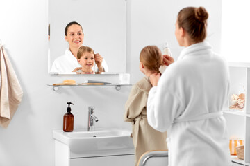 Poster - beauty, family and people concept - happy smiling mother and daughter looking to mirror and braiding hair in bathroom