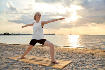 Wall Mural - fitness, sport, and healthy lifestyle concept - happy woman doing yoga warrior pose on beach over sunset