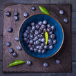 Blueberries with green leaves in a bowl, on a stone stand, on a dark blue wooden table. Healthy food. Diet. Top view.