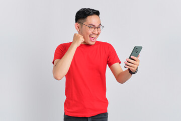 Wall Mural - Excited young Asian man looking at mobile phone screen gesturing yes with raised fist reacting to online news isolated over white background