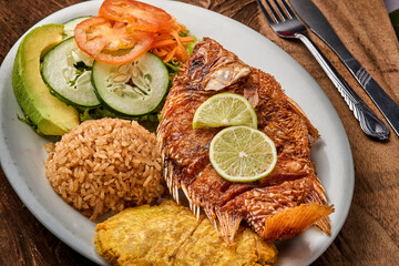 foto horizontal de pescado frito con arroz de coco y patacones y ensalada verde en una mesa de madera con cubiertos