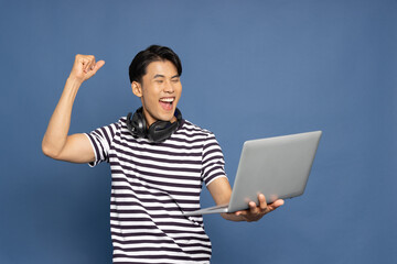 Happy young Asian pro gamer with headphones holding computer laptop and win in online video game isolated over blue background, Cyber sportsman playing tournaments concept