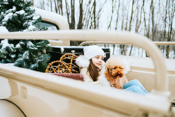 cute beautiful little Armenian girl in winter coat and knitted hat and pet poodle dog sit near beige retro pickup truck decorated for Christmas and New Year vintage interior suitcases, skis, sledge