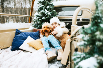 cute beautiful little Armenian girl in winter coat and knitted hat and pet poodle dog sit near beige retro pickup truck decorated for Christmas and New Year vintage interior suitcases, skis, sledge