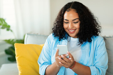 Wall Mural - cheerful african american woman using smartphone sitting at home