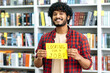 Looking for a job. Smiling unemployed indian guy, stands in the library against background of bookshelves, holds a sign in his hand that says looking for a job, looks at camera, hopes to get a job
