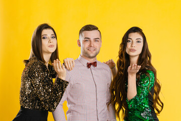 Young adult girls hug a guy and make an air kiss to the camera. Festive corporate party of friends. A man with two girls on a yellow background in festive clothes look at the camera