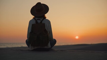 Wall Mural - Young girl in a hat sitting on the beach and looking at the sunset