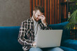 © ViDi Studio - Young sad tired man he wears brown shirt hold use work on laptop pc computer keep eyes closed rub put hand on nose sitting on blue sofa in own living room apartment stay home indoor flat on weekends