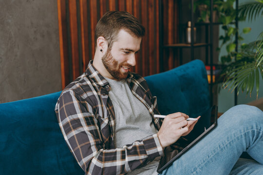 Young happy graphic designer man he wears shirt hold in hand work use write draw stylus pc pen on tablet pc computer sit on blue sofa in own living room apartment stay home indoor flat on weekends