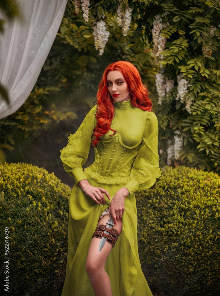 Fantasy red-haired dangerous woman medieval assassin queen with dagger in  sheath on leg leather belt. Green vintage old style dress. Girl princess  warrior. Garden summer nature trees. Red long hair Stock-foto |