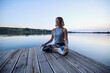 A calm yogi woman in a lotus position is meditating on the dock.