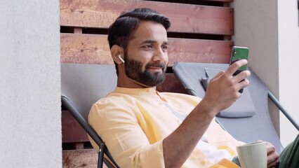 Wall Mural - Relaxed indian man sitting outdoor house on chair chilling, drinking coffee, wearing earbud using smartphone looking at cell phone watching video news, checking social media, listening music podcast.