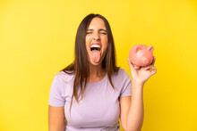 Hispanic Young Adult Woman With Cheerful And Rebellious Attitude, Joking And Sticking Tongue Out. Piggy Bank Savings Concept
