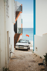 Wall Mural - Alley with an old white car and the ocean at the background. Algarve, Portugal