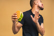 © denis_vermenko - Stop sign, bad smell. Young african american black man eating hamburger isolated over beige background. Diet concept.
