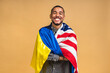 © denis_vermenko - Portrait of positive african american black guy holding usa and ukrainian flags, isolated over beige background.