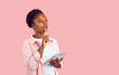 Beautiful Afro American woman in casual shirt standing on blank pink background, holding notebook, looking away, biting pencil and thinking. Female student or copywriter writing ideas for new article