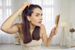Portrait of woman in her 20s or 30s looking at her reflection with scared nervous expression as she notices bad signs like scalp dandruff, hair thinning, or hair falling out. Hair loss problem concept