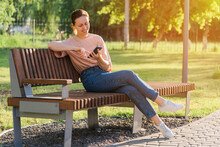 Beautiful Brunette With A Mobile Phone Sits On A Bench In The Park