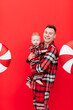 The concept of parenthood. Smiling young father is holding and hugging his baby son in the same pyjamas on red background with Christmas big lollipops, dad enjoying time with baby boy. Fathers Day