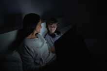 Smiling Woman And Boy In Sleepwear Lying On Bed With Laptop While Watching Movie At Night And Looking At Each Other