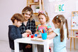© Татьяна Волкова - Young cheerful woman sitting at table around children. Teacher explaining pupil boy how to complete task in classroom.